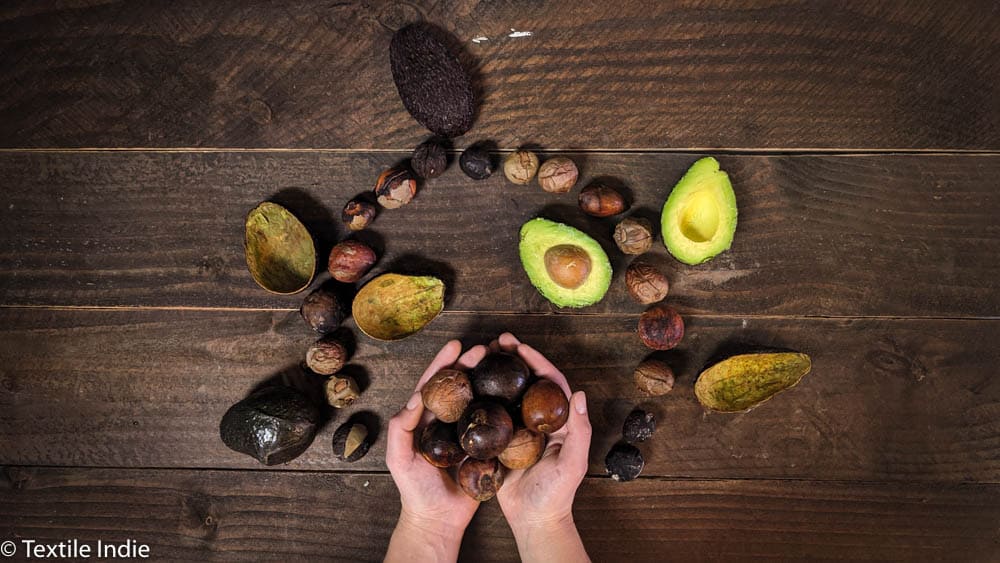 Avocado pits and skin for dyeing are arranged on a wooden table, a woman's hands hold a pile of avocado pits