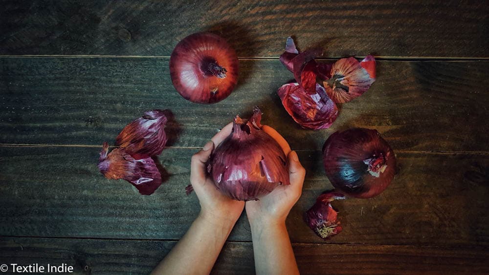 a woman holding a red onion, ready for natural dyeing. Red onions and onion skins are artfully arranged on a table 
