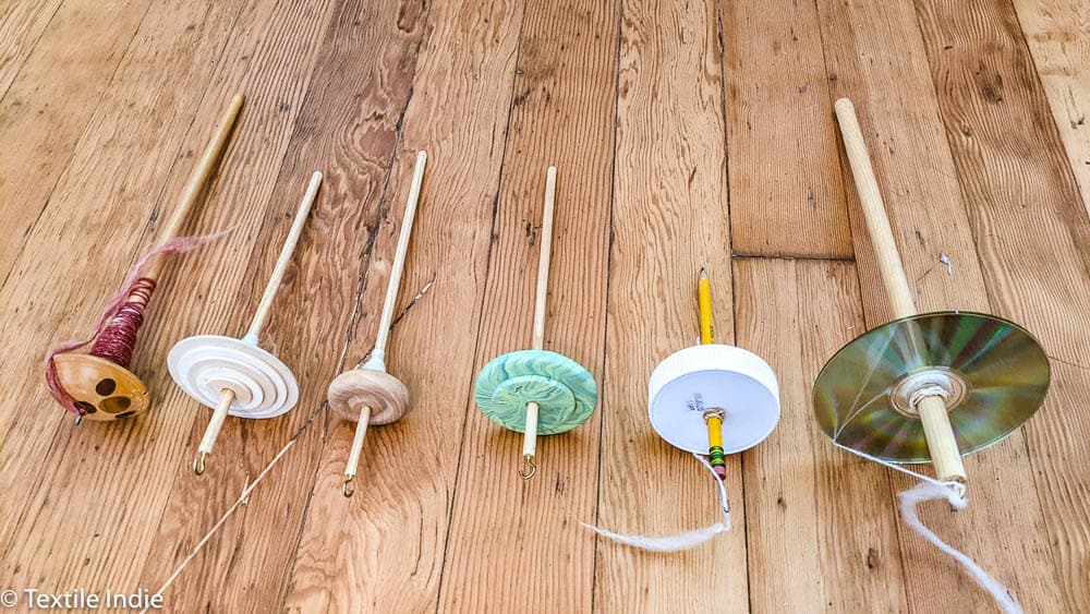 a row of drop spindles laid out on a wood floor, the first is store-bought, the five next are homemade 