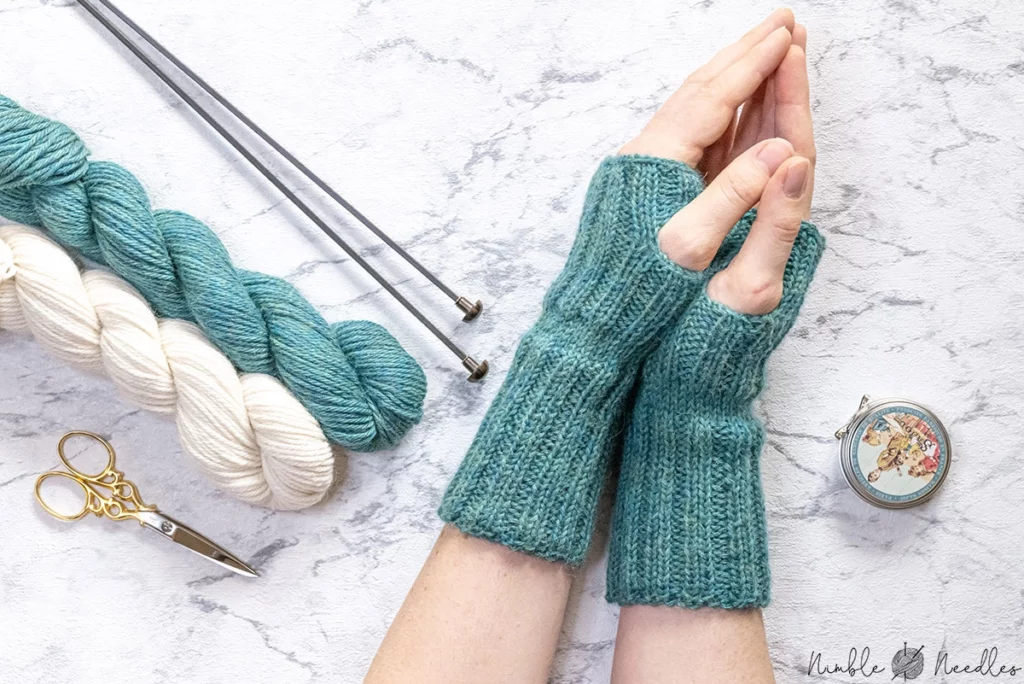 Teal green fingerless gloves on a pair of womens hands, knitting needles and skeins of yarn in the background