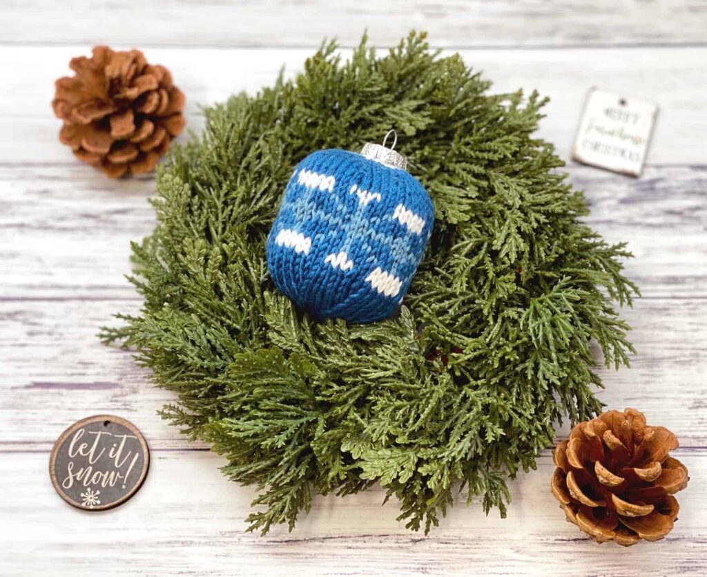 knit Christmas ball in a bed of evergreens with pinecones in the background