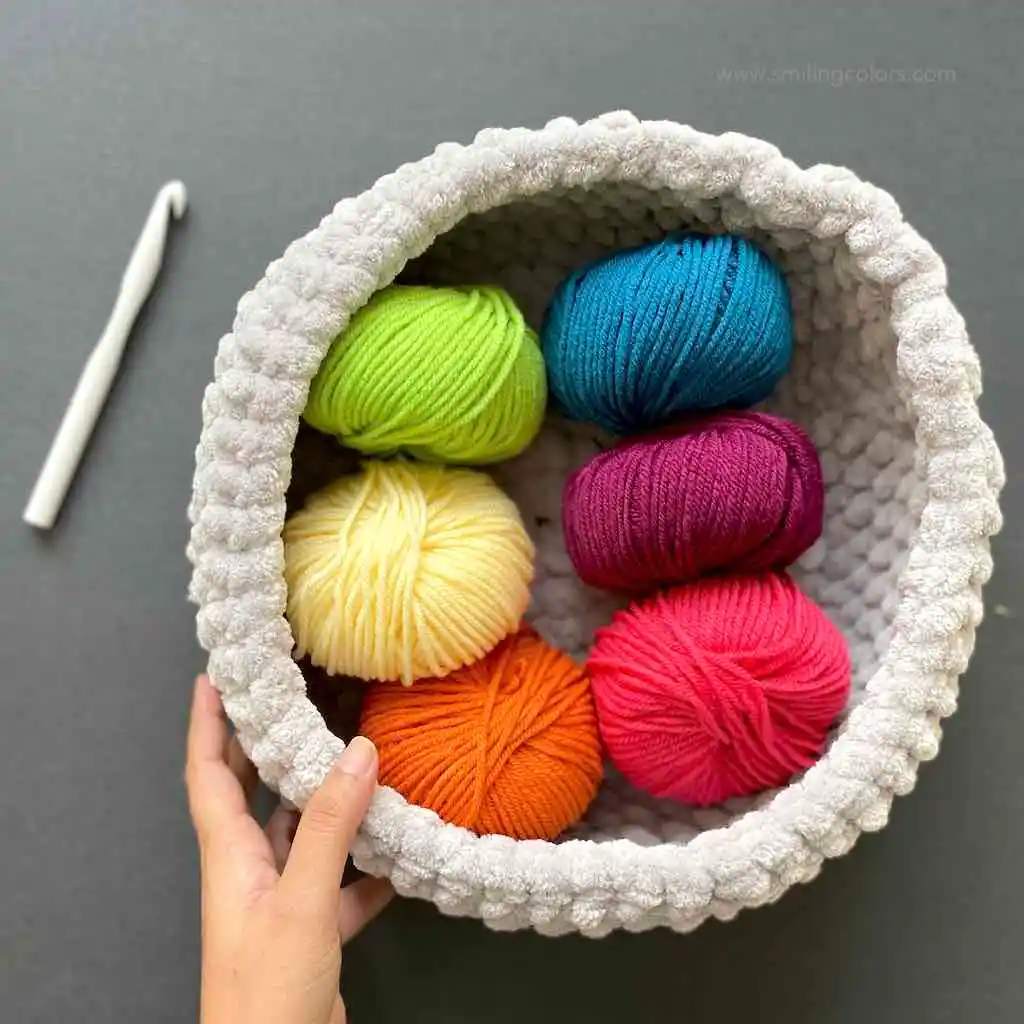 Soft crochet basket in white, holding 6 balls of brightly colored yarn, white crochet hook sits to the side
