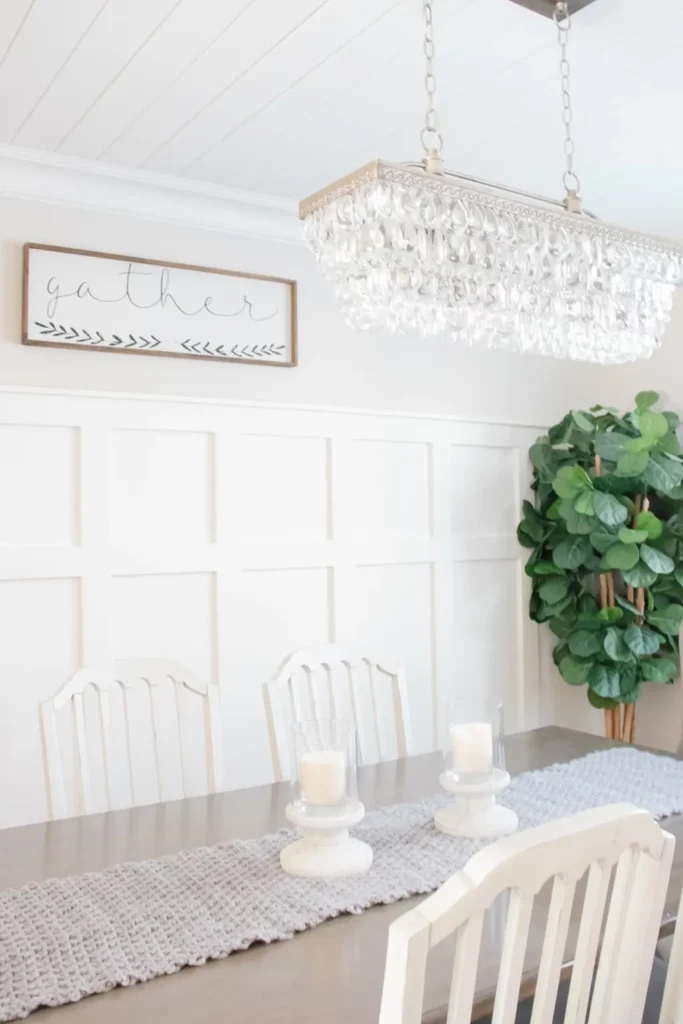 white knit table runner on a farmhouse table in a dining room lined with white paneling, a plant is in the background, and a crystal chandelier hangs from the ceiling