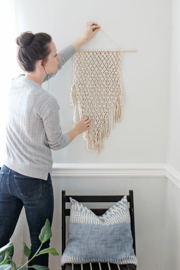 A woman in a grey sweater and blue jeans hangs a delicately knotted macrame wall hanging 