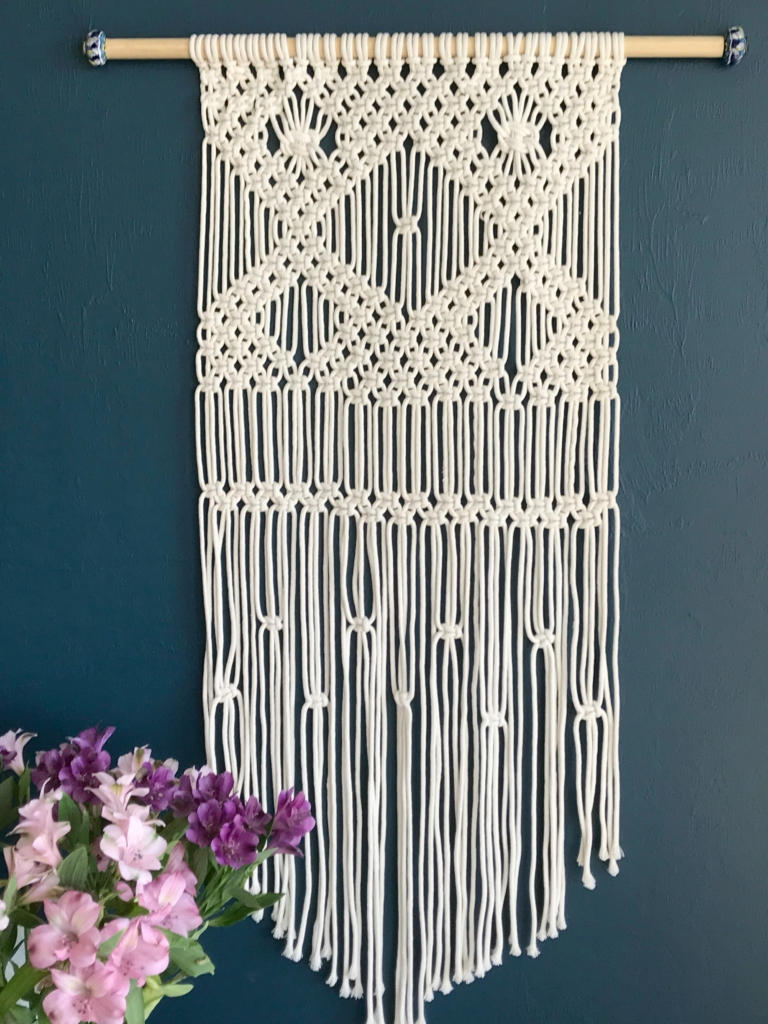 a large natural color macrame wall hanging hangs from a dowel against a teal wall 