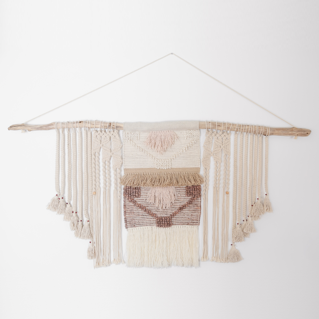 A large textured macrame wall hanging in various cream hues is hanging from a long piece of driftwood against a white wall