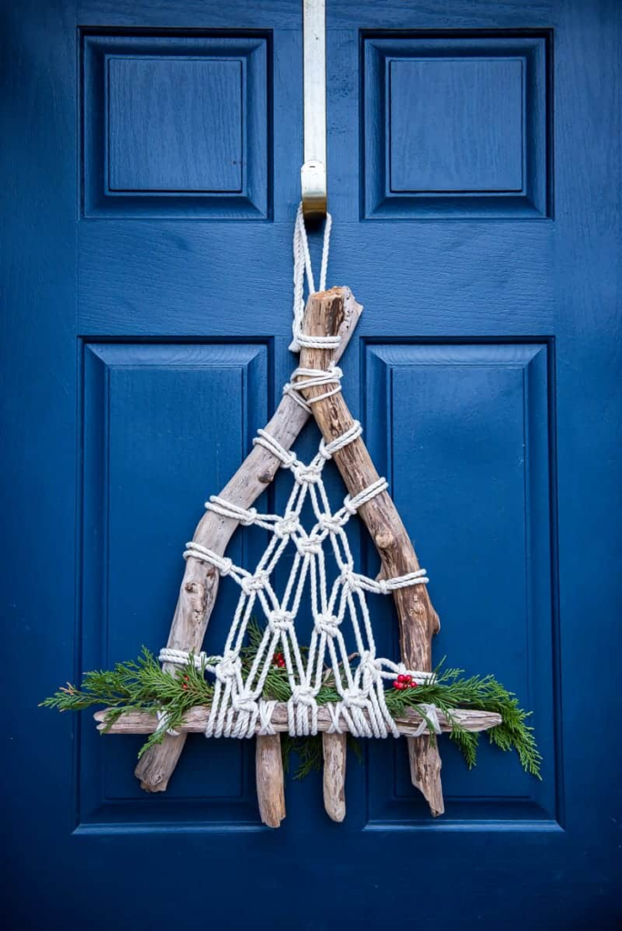A Christmas style macrame hanging sits on a bright blue door. The frame of the hanging is made with driftwood and there's greenery and berries tucked in