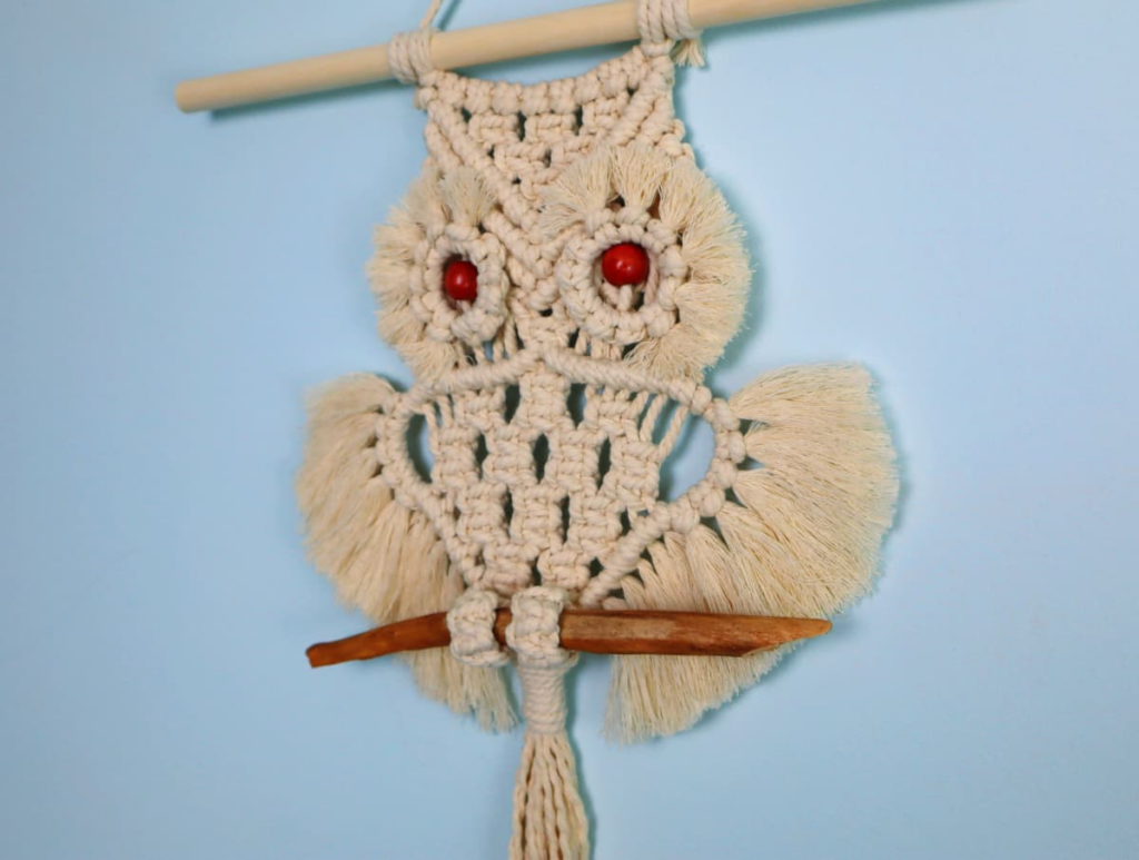 An owl macrame wall hanging with red bead eyes. It's hanging from a dowel against a blue wall.