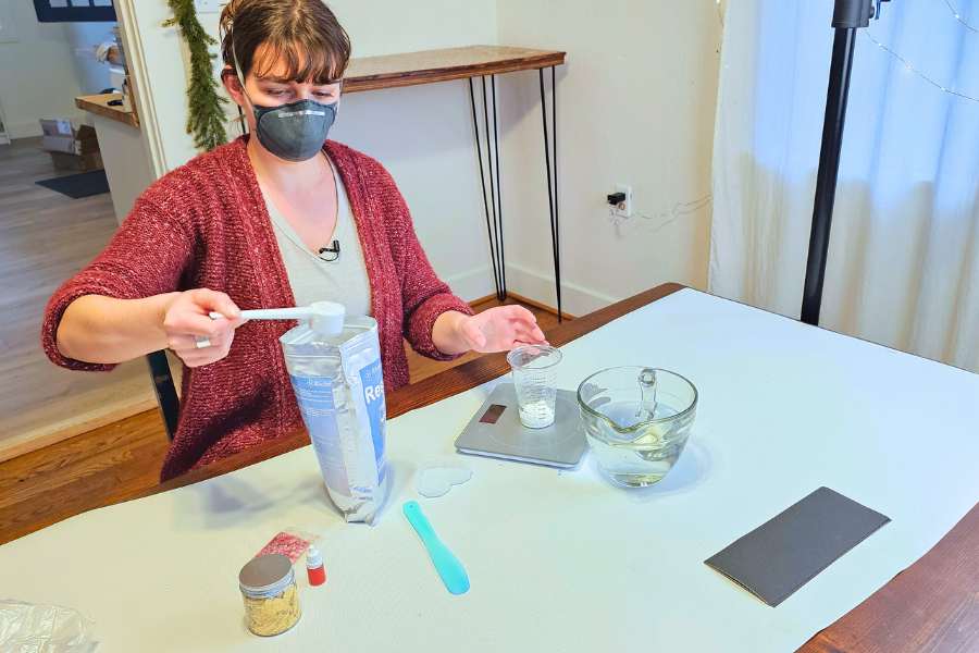 Brittany in a mask measuring out the resin base into a plastic cup which is sitting on a digital scale