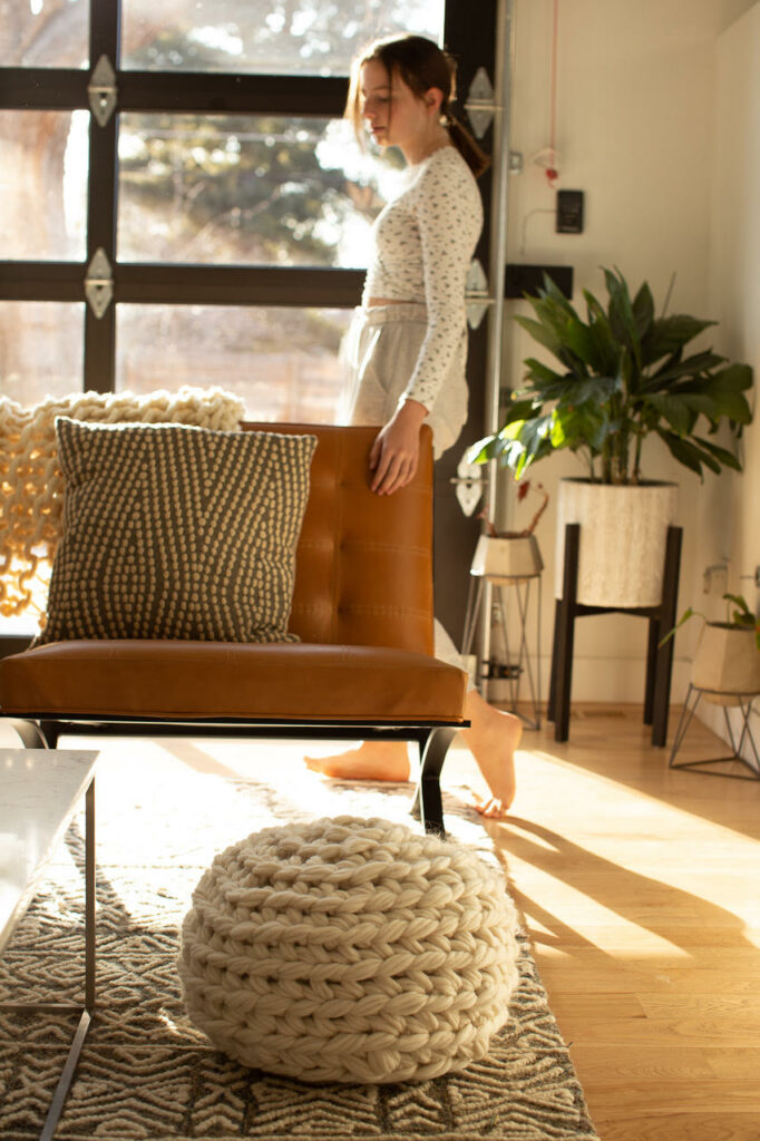 Flax and Twine crochet pouf next to a brown leather couch, a woman is standing behind the couch