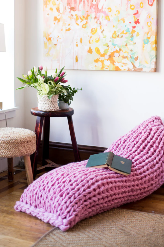 a bright pink body pillow leaning against the wall, the pillow uses the arm-knitting technique 