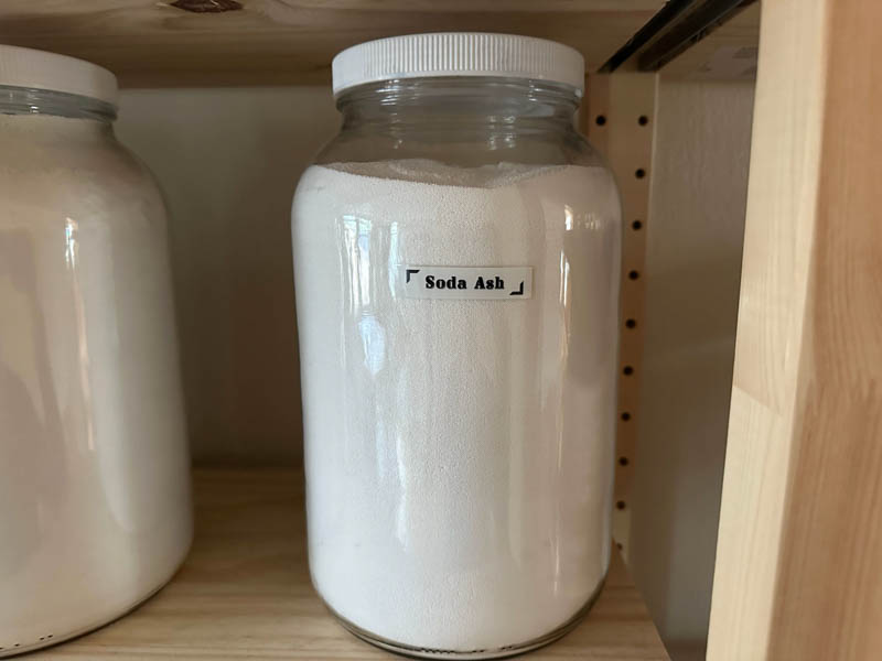 Large gallon size jar with the label "soda ash" filled with soda ash.