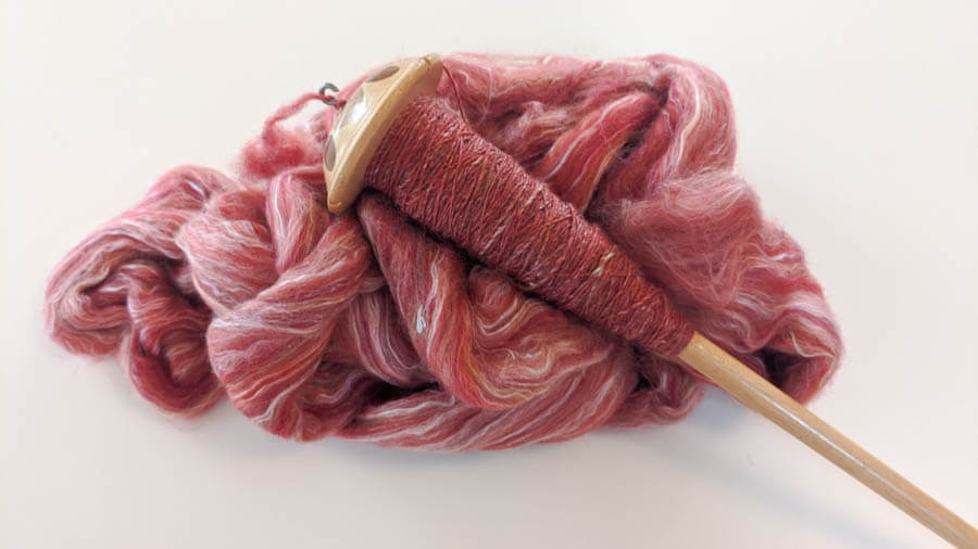 A drop spindle wound with bright pink yarn, sitting on a pile of brightly dyed wool roving