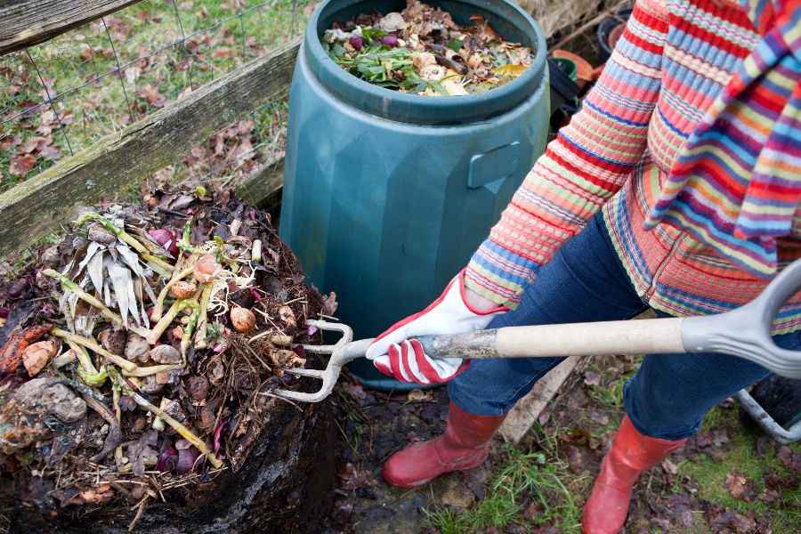 A woman in a bright sweater and pink rainboots turning over compost with a garden fork 